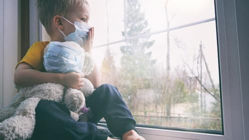 A young boy wearing a surgical face mask and holding a teddy bear looking out a window
