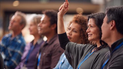 A woman at a conference, raising her hand to ask a question.