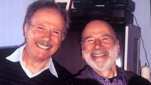 Two smiling men with some equipment and a framed photo behind them.