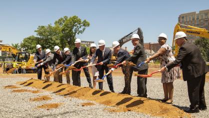 Men and women in hard hats breaking ground with shovels. Construction vehicles in the background.