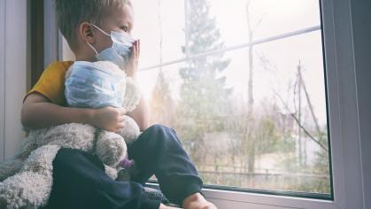 A young boy wearing a surgical face mask and holding a teddy bear looking out a window