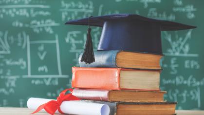 A graduation cap on top of a pie of books next to a diploma with a chalkboard in the background