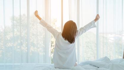 Photo of young happy woman in pajama stretching her arms and smiling while sitting on bed after sleep