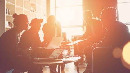 Silhouette of startup business team meeting on the couch in a big open space office. Five people. Intentional sun glare and lens flares. Wide screen, panoramic