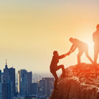 Cityscape in background with people at the top of a rock, one helping another climb to the top