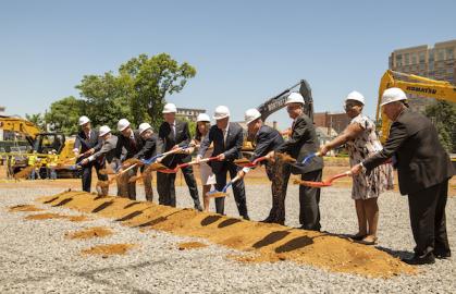 Men and women in hard hats breaking ground with shovels. Construction vehicles in the background.