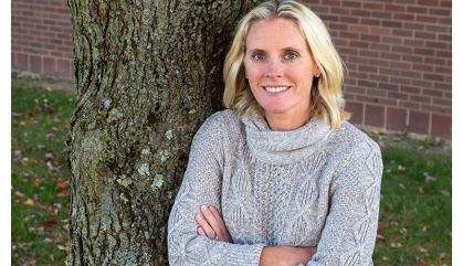 Woman in a grey turtleneck standing by a tree crossing her arms.